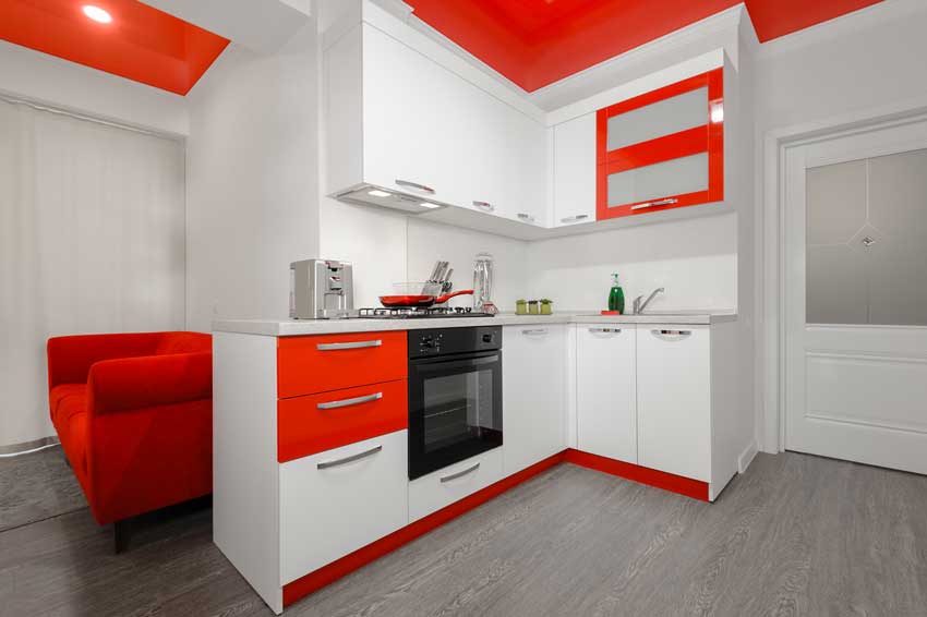 Piccola cucina ad angolo bianca con cassetti e zoccolatura rossi, pareti bianche e soffitto rosso.