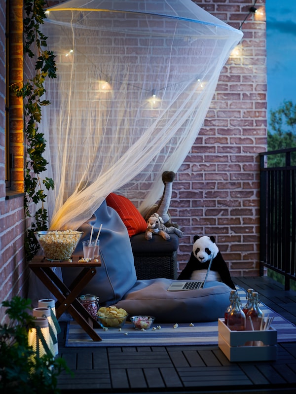 Angolo relax su un balcone con pareti in mattoni, allestito per una serata cinema, con rete appesa, cuscini e peluche - IKEA