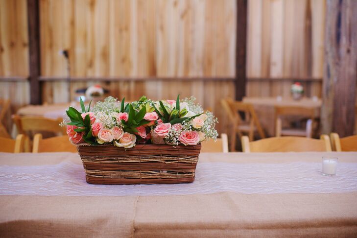 Il tavolo di testa era coperto di tela con un corridore da tavolo di pizzo. Cesti di legno e vimini di fiori bianchi e rosa con vegetazione ammorbidivano l'impostazione rustica della tavola.