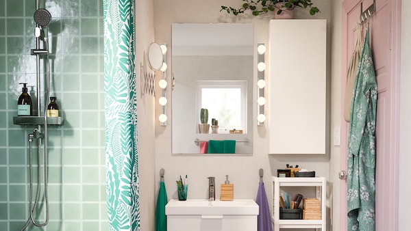 Una doccia piastrellata di verde accanto a una stanza rosa con un lavabo bianco e un mobile a parete, accanto a due lampade da parete a LED LEDSJÖ accese - IKEA